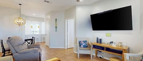 A living room with a ceiling fan, a flat-screen TV mounted on the wall, a grey sofa, a wooden coffee table, and a patterned rug.