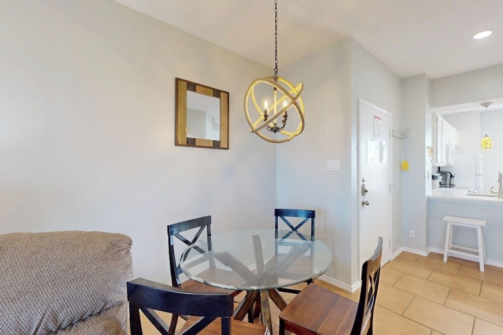 A dining area with a round glass table, four chairs, a hanging light fixture, and a kitchen visible in the background.