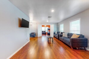 A spacious living room with hardwood flooring, a flat-screen TV mounted on the wall, a dark-colored sofa, and a coffee table. - The Seahorse - Sleeps 12, Near Beach & Downtown (Galveston)