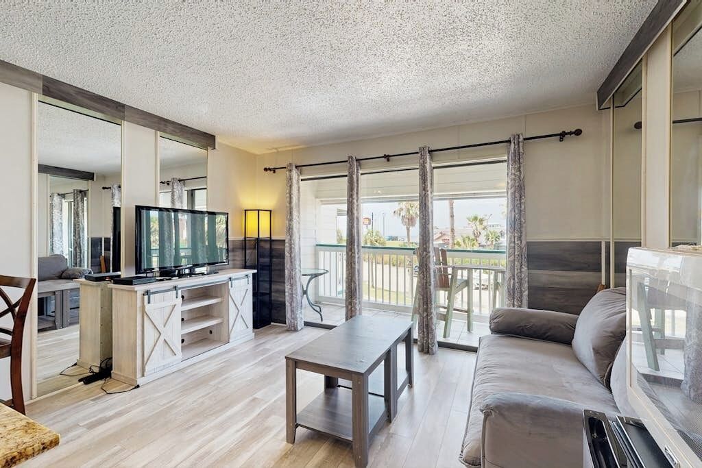 A modern living room with a flat-screen TV, a sofa, a coffee table, and a balcony with curtains.