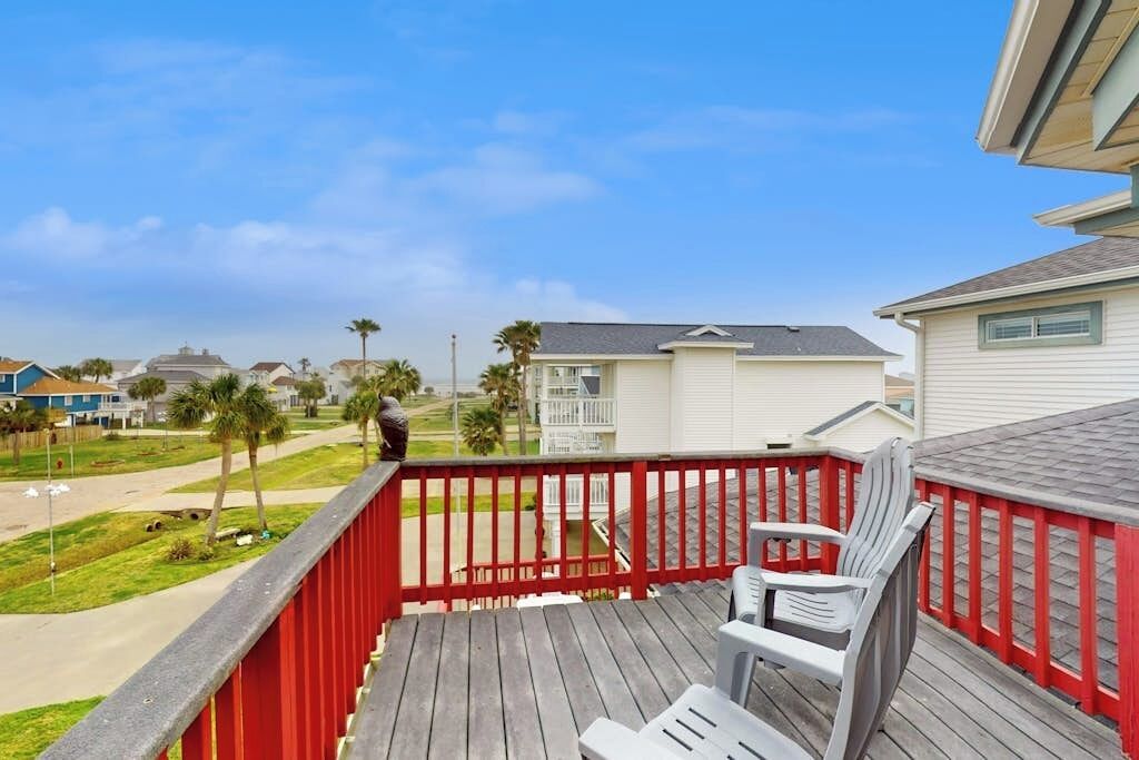 A wooden deck with a red railing, two white chairs, and a view of a residential area with houses and palm trees.