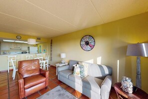 Ein Wohnzimmer mit einer Couch, einem braunen Ledersessel, einem Essbereich mit Tisch und Stühlen, einer Küche im Hintergrund und einer Wanduhr.