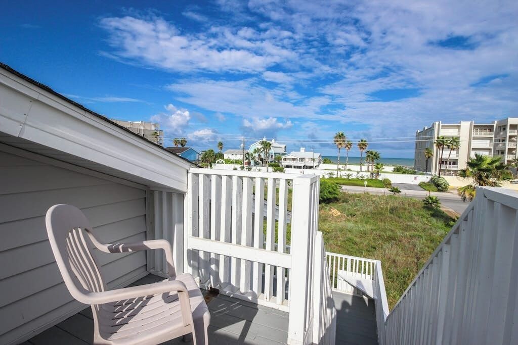 A balcony with a white chair, a white railing, and a view of the beach and buildings.