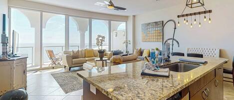 A spacious living area with a large island kitchen, a ceiling fan, and a view through large windows.