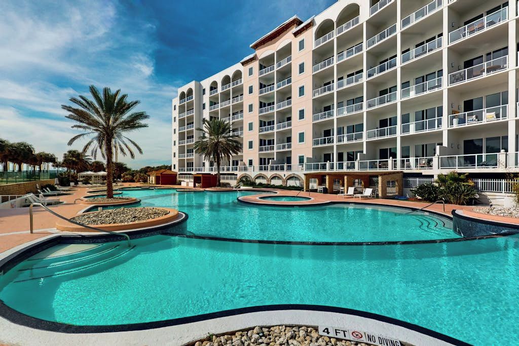 A hotel with a large swimming pool, multiple balconies, and palm trees.