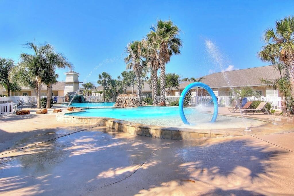Une piscine agrémentée d’un élément décoratif aquatique, bordée de palmiers, avec un bâtiment visible au loin.