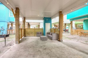 An outdoor covered patio area with wooden furniture, a concrete floor, and a view of a building with blue siding. - Stack Overflow - Balcony, Pool & Dog-Friendly (Port Aransas)