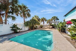 A swimming pool with a clear blue water, surrounded by palm trees and a white building with a red awning. - Coastal Hideaway - Screened Porch, Pool & Dogs OK (Port Aransas)