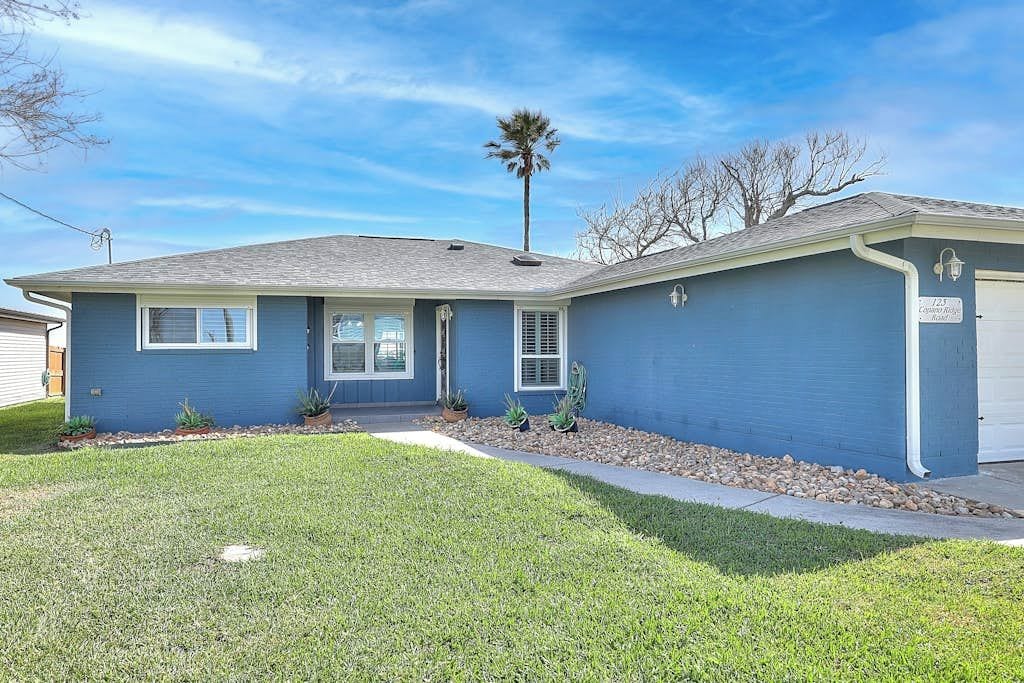 A single-story residential house with a blue exterior, a front door, and a small front yard with a palm tree and potted plants.