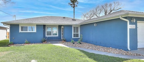 A single-story residential house with a blue exterior, a front door, and a small front yard with a palm tree and potted plants.