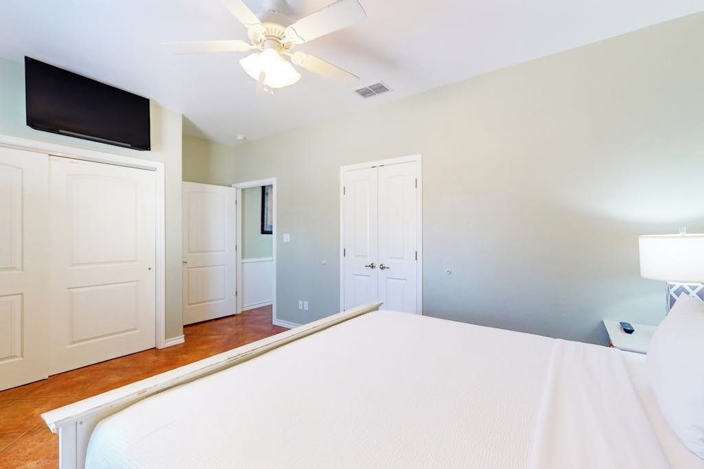 A bedroom with a bed, a ceiling fan, a TV mounted above the bed, and a nightstand with a lamp.