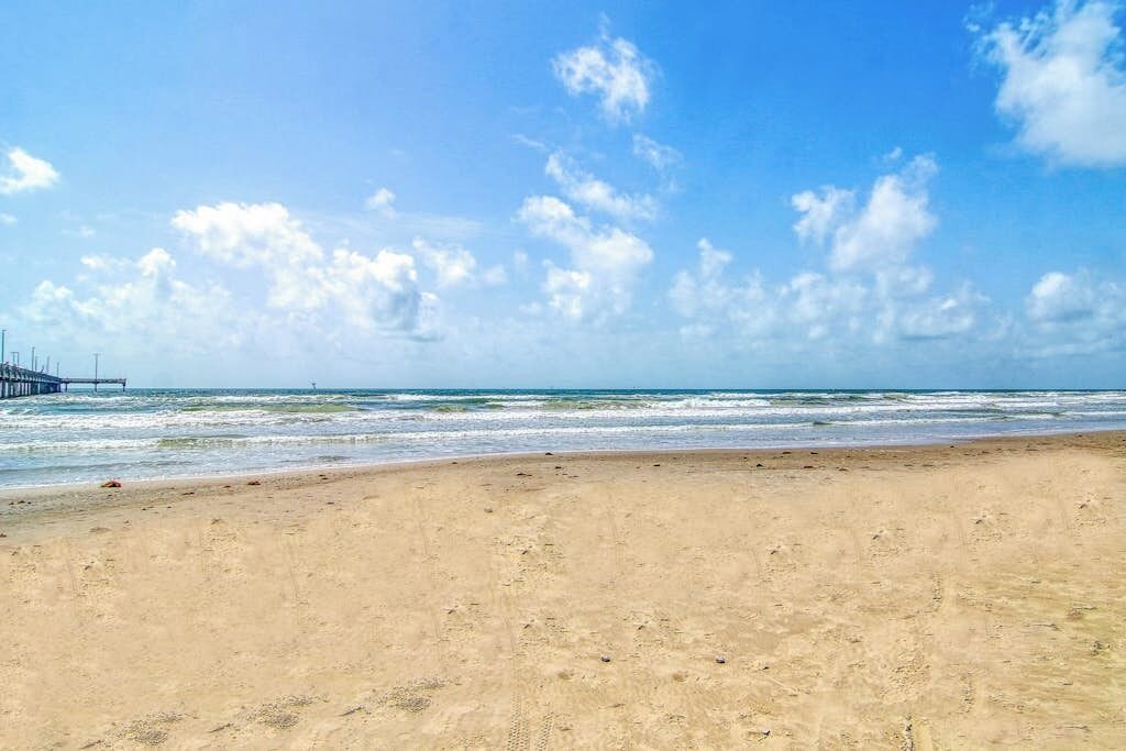 A beach with a pier extending into the water, sandy shore, and a clear sky with clouds.