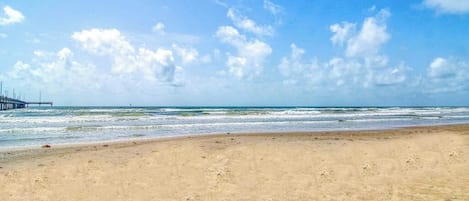 A beach with a pier extending into the water, sandy shore, and a clear sky with clouds.