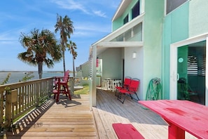 A wooden deck with outdoor furniture, a view of the water, and a building with a green facade. - Puente Vista 133 - Bay-View Deck & Shared Pool (Corpus Christi)