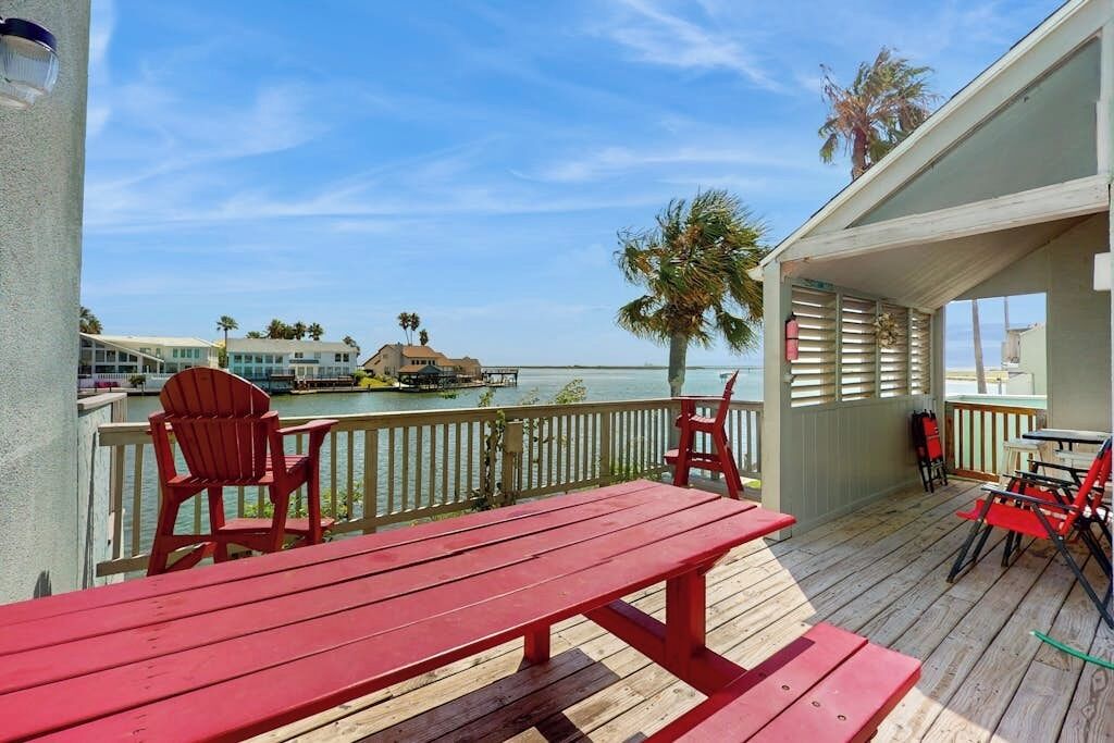Eine Holzdar Terrasse mit roten Stühlen und einem Tisch, mit Blick auf ein Gewässer, in dessen Ferne ein Boot und Gebäude liegen.