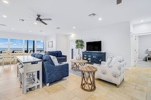 A modern living room with a dining area, a ceiling fan, a television, and a view of the ocean. - Bay Breeze Beauty - Waterfront, Hot Tub & Dogs OK (Rockport)