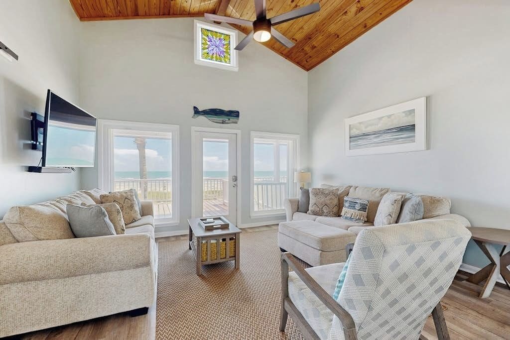 A spacious living room with a high ceiling, a ceiling fan, a television mounted on the wall, and a large window overlooking the ocean.