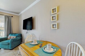 A room with a sofa, a dining table with chairs, a TV mounted on the wall, and a window with curtains. - Beachside Escape - Gulf-Front Condo with Pool (Galveston)