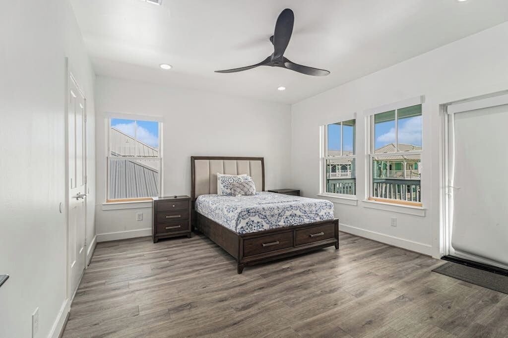A bedroom with a bed, nightstands, a ceiling fan, and windows with a view.