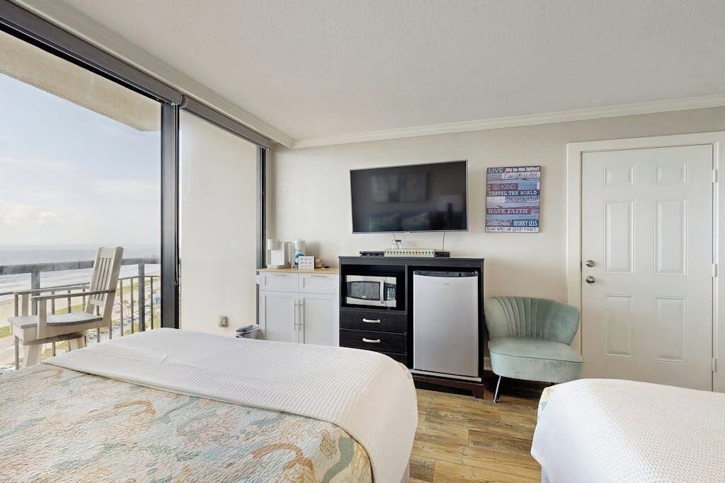 A hotel room with a bed, a desk with a television, a small refrigerator, and a view of the ocean.