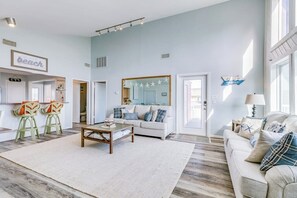 Ein geräumiges Wohnzimmer mit einem weißen Sofa, einem Couchtisch und einem großen Spiegel. Zwei Sessel mit Kissen, ein Teppich und ein an der Wand befestigtes Schild mit der Aufschrift 'beach'.