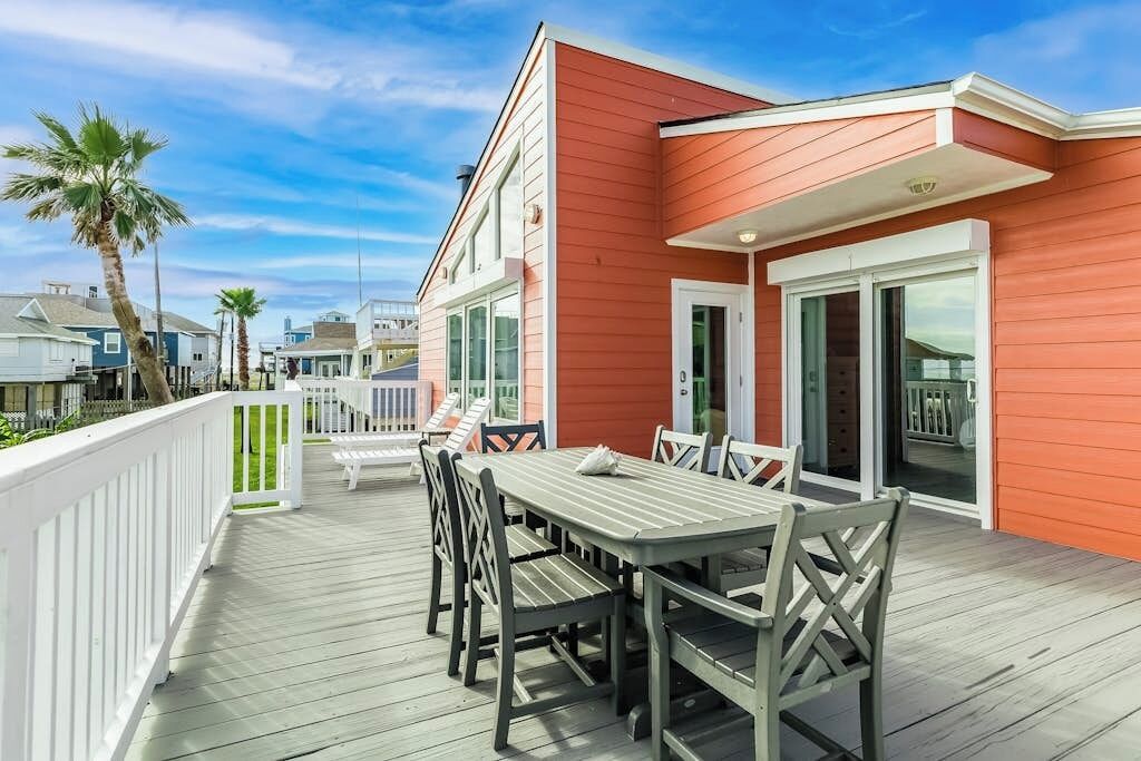 A covered outdoor deck with a dining table and chairs, a white fence, and a red building in the background.