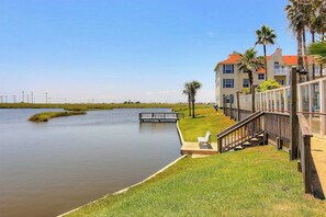 A waterfront property with a grassy area, a wooden deck, and a building with a red roof. - Beach Club - Ocean Breeze Retreat – Island Condo (Corpus Christi)