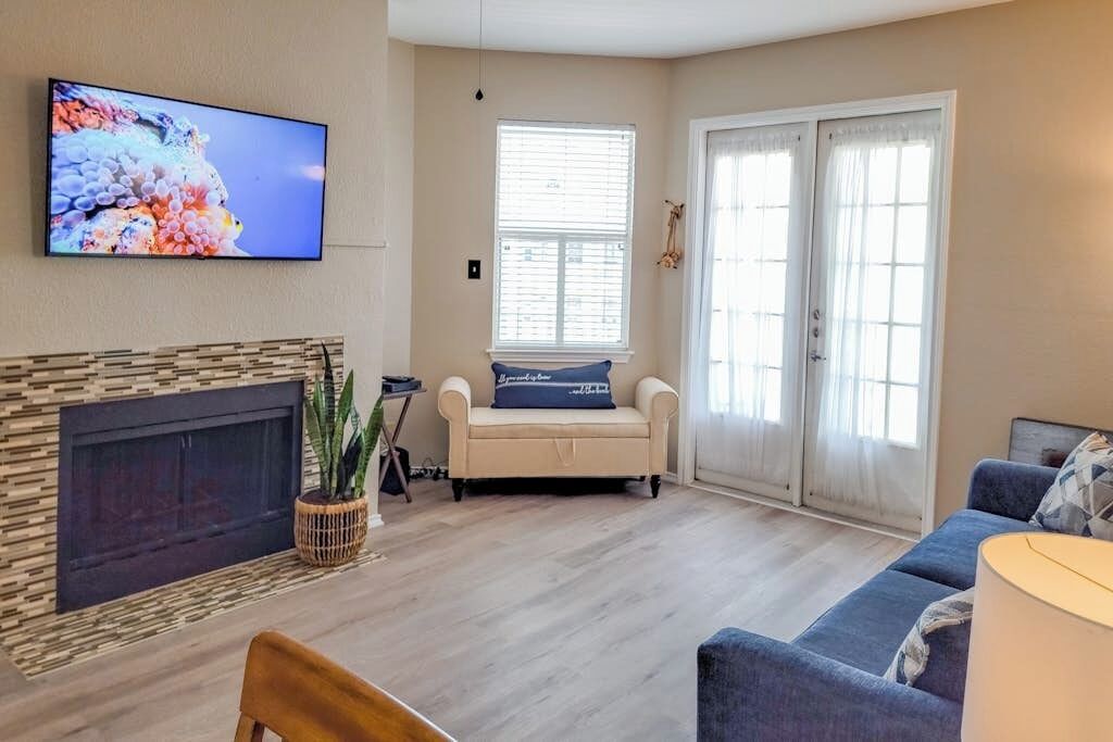 Een woonkamer met een open haard, een aan de muur bevestigde televisie en een groot raam met zonwering.