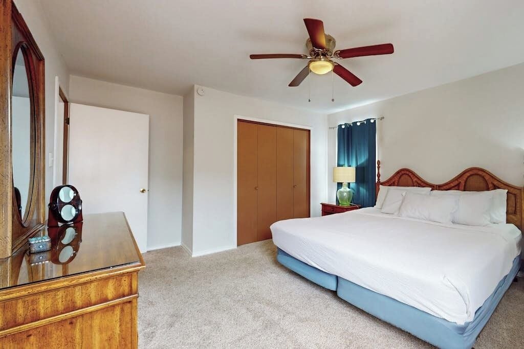 Une chambre à coucher comprenant un lit, une commode en bois, un ventilateur de plafond, une porte et une table de chevet avec une lampe.