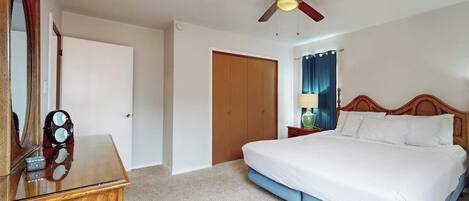 A bedroom with a bed, a wooden dresser, a ceiling fan, a door, and a nightstand with a lamp.