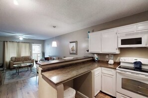 A compact kitchen with white cabinets, a microwave, and a stove. There is a dining area with a table and chairs, and a sofa. - Las Palmas Condominiums #10 (South Padre Island)