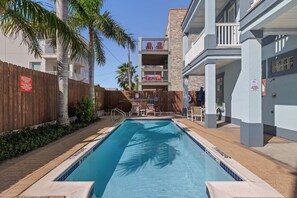 A swimming pool with a wooden fence, palm trees, and a building with balconies. - Retama Landing #3 - Pool & Balcony, Walk to Beach (South Padre Island)