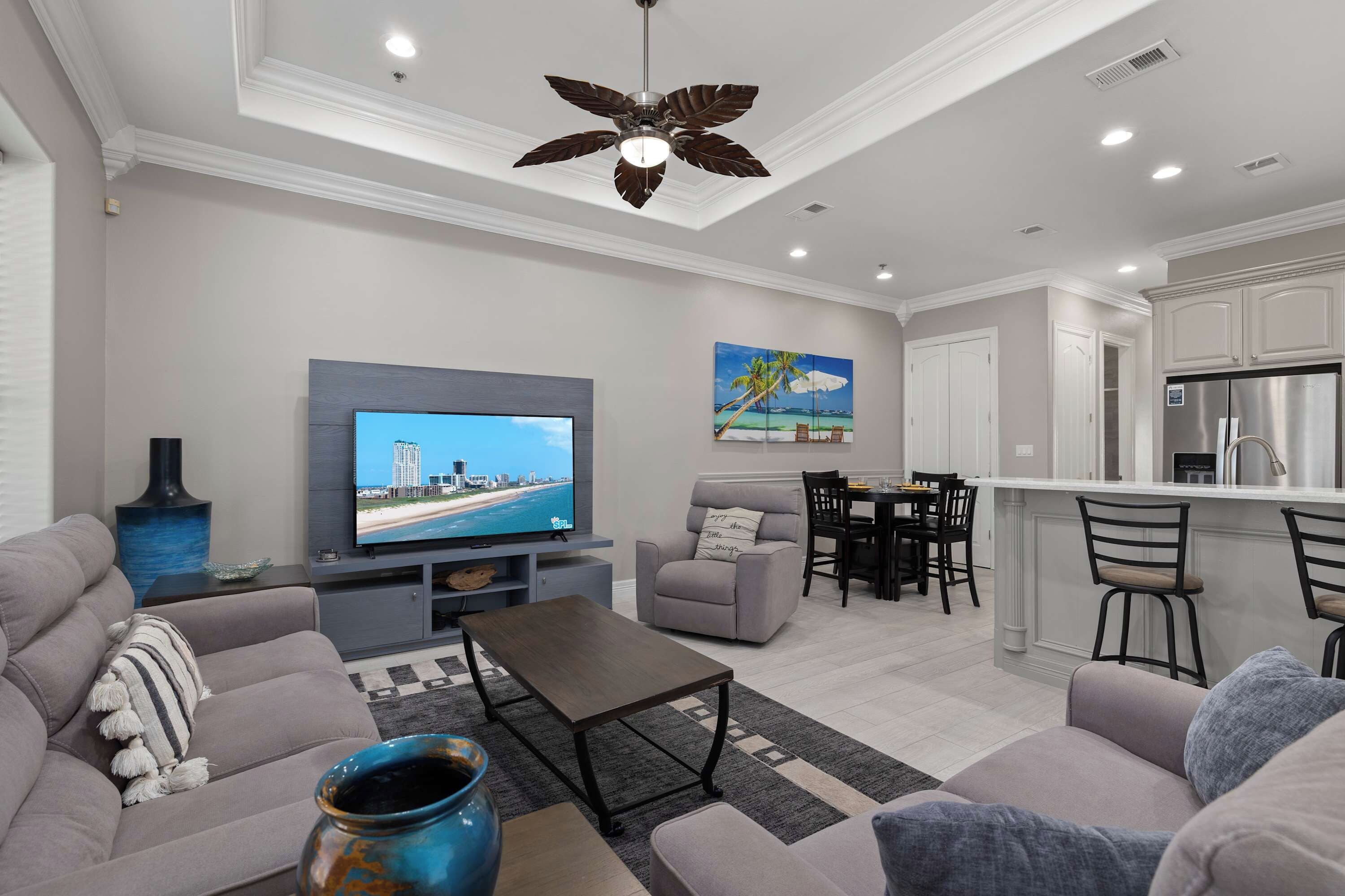 Ein modernes Wohnzimmer mit Flachbildfernseher, grauen Sofas, einem Couchtisch und einem Essbereich, im Hintergrund ist eine Küche zu sehen.