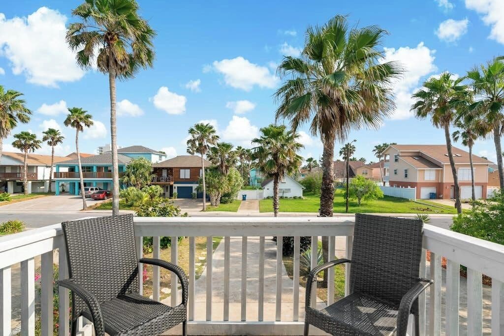 Een balkon met rieten stoelen, uitkijkend op een woonstraat met palmbomen en huizen.