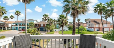 Een balkon met rieten stoelen, uitkijkend op een woonstraat met palmbomen en huizen.
