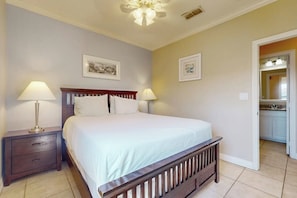 A bedroom with a bed, two bedside tables, and a nightstand. There is a door leading to a kitchen. - Garden Breeze Condominiums #3 (South Padre Island)