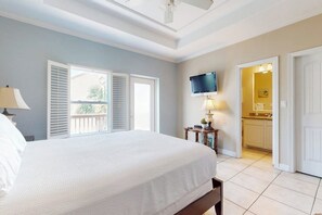 A bedroom with a bed, a television, a window with blinds, and a door leading to a bathroom. - Garden Breeze Condominiums #3 (South Padre Island)