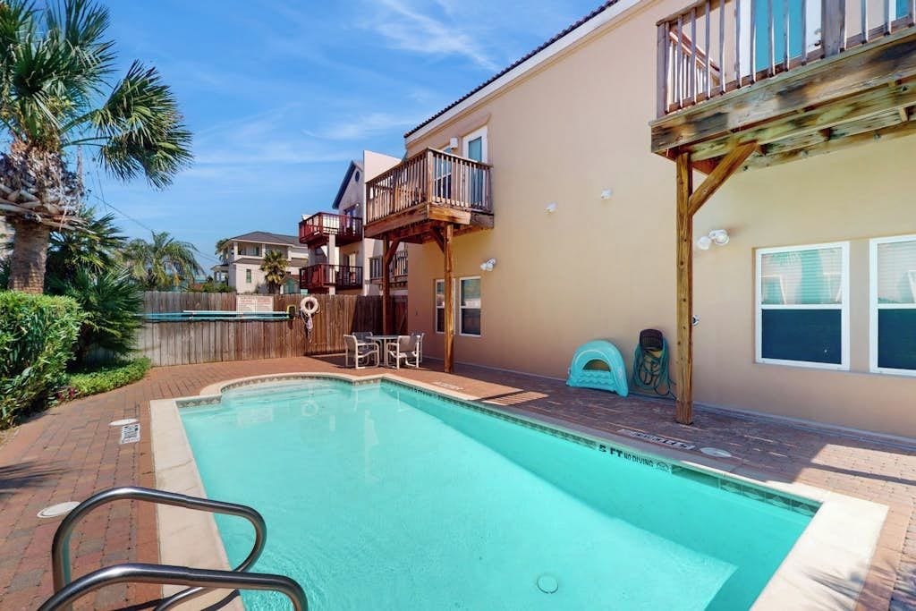 A swimming pool with a wooden deck, a covered outdoor seating area, and a two-story building with balconies.