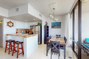 A modern kitchen with a central island, bar stools, a dining table with chairs, and a cityscape painting on the wall. - Sunchase IV #316 - Heated Pool & Ocean Views (South Padre Island)