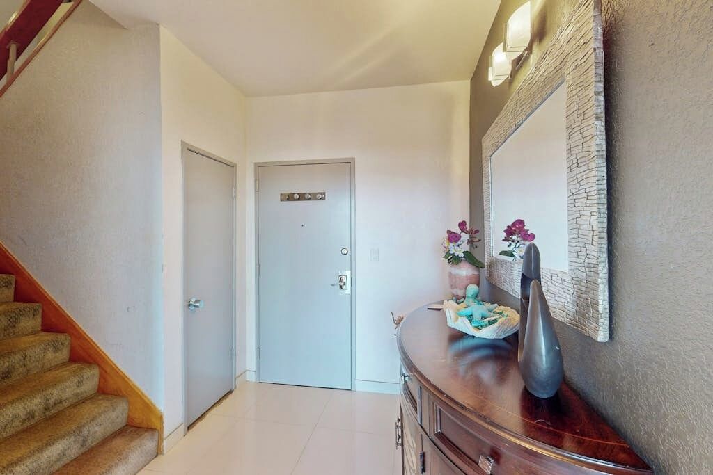 Un pasillo con una escalera de madera a la izquierda, una puerta cerrada al final y una pared decorativa con un jarrón y flores.