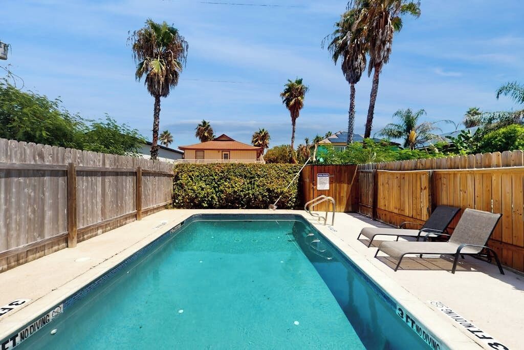 A rectangular swimming pool with a wooden fence and lounge chairs on one side.