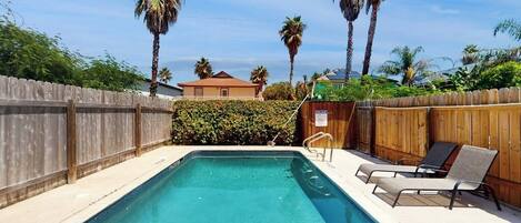 A rectangular swimming pool with a wooden fence and lounge chairs on one side.