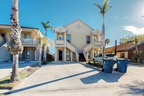 A two-story residential building with a central entrance, a covered driveway, and a parking area with two cars and trash bins. - Saturn Serenity 1 - Shared Pool & Dog-Friendly (South Padre Island)