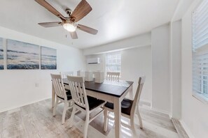 A dining area with a wooden table and chairs, a ceiling fan, and artwork on the wall. - Lazy Seagull Beach House - Downstairs (South Padre Island)