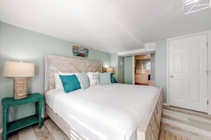 A bedroom with a bed, bedside tables, a door, and a hallway leading to a bathroom. - Lazy Seagull Beach House - Downstairs (South Padre Island)