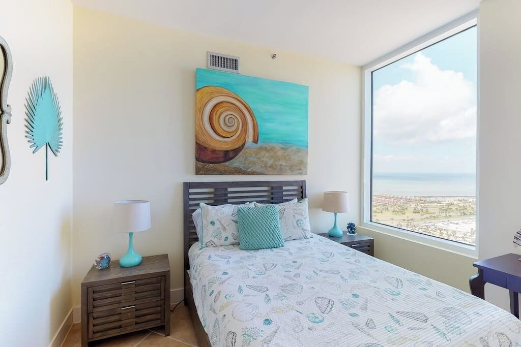 Un dormitorio con cama, mesitas de noche, un cuadro y una ventana con vistas.