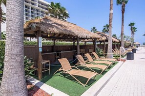Un área junto a la piscina con sillones de descanso, una estructura de techo de paja y un edificio al fondo.