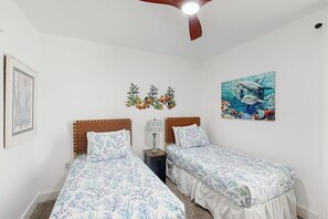Een slaapkamer met twee bedden, een plafondventilator, een aan de muur bevestigde lamp en twee ingelijste kunstwerken.