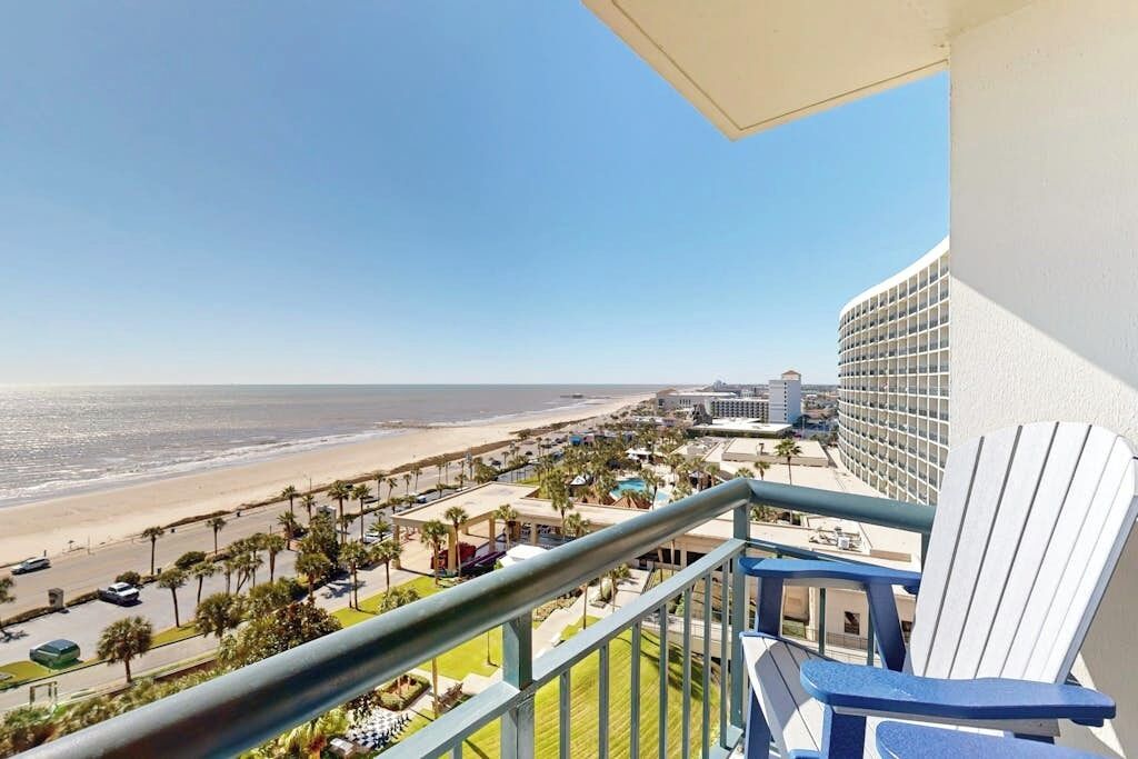 A balcony with a view of a beach and ocean, a chair, and a building with a curved facade.