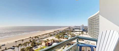 A balcony with a view of a beach and ocean, a chair, and a building with a curved facade.